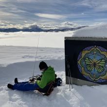Me, seated in the snow, logging, with low clouds and mountains in the background