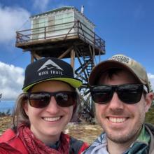 Man and woman take selfie in front of fire lookout tower. 